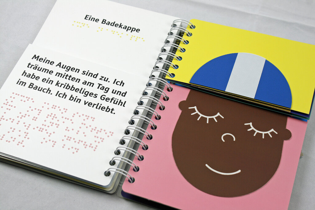 Aufgeschlagenes Ringbuch mit geteilten Seiten zum Klappen, links der Text in Braillelack und Großdruck, rechts Gesicht mit geschlossenen Augen, Badekappe
