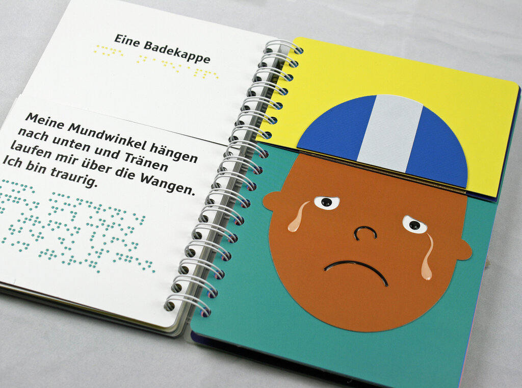Aufgeschlagenes Ringbuch mit geteilten Seiten zum Klappen, links der Text in Braillelack und Großdruck, rechts Gesicht mit Tränen, Badekappe