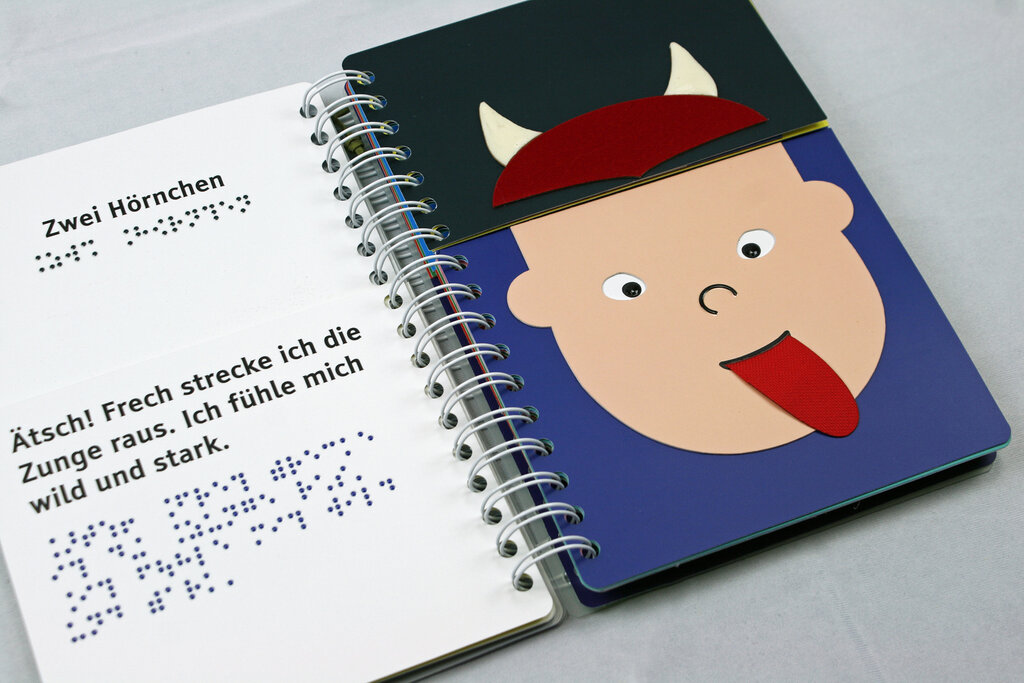 Aufgeschlagenes Ringbuch mit geteilten Seiten zum Klappen, links der Text in Braillelack und Großdruck, rechts Gesicht mit ausgestreckter Zunge, zwei Hörnchen 