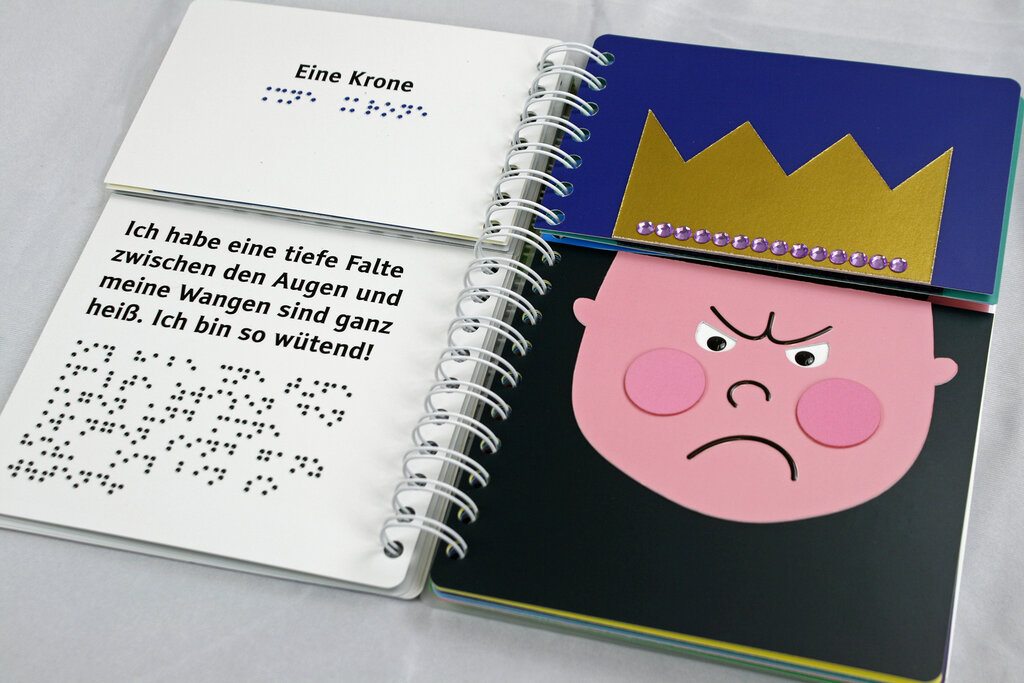 Aufgeschlagenes Ringbuch mit geteilten Seiten zum Klappen, links der Text in Braillelack und Großdruck, rechts Gesicht mit tiefer Falte zwischen den Augen, Krone 