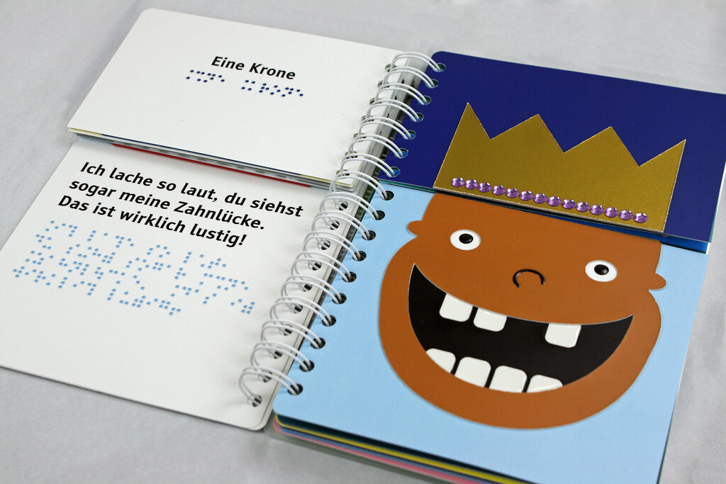 Aufgeschlagenes Ringbuch mit geteilten Seiten zum Klappen, links der Text in Braillelack und Großdruck, rechts lachendes Gesicht mit Zahnlücke, Krone