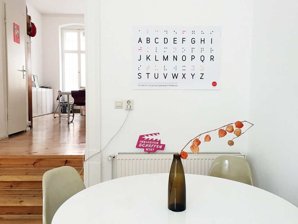 Im Vordergrund ein runder Tisch mit zwei Stühlen, dahinter an der Wand hängt ein Plakat mit dem Alphabet in Brailleschrift.
