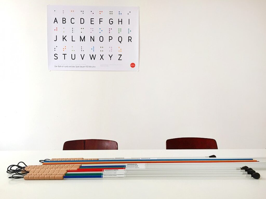 Im Vordergrund ein Tisch, auf dem mehrere Blindenstöcke liegen. Dahinter an der Wand hängt ein Plakat mit dem Alphabet in Brailleschrift.