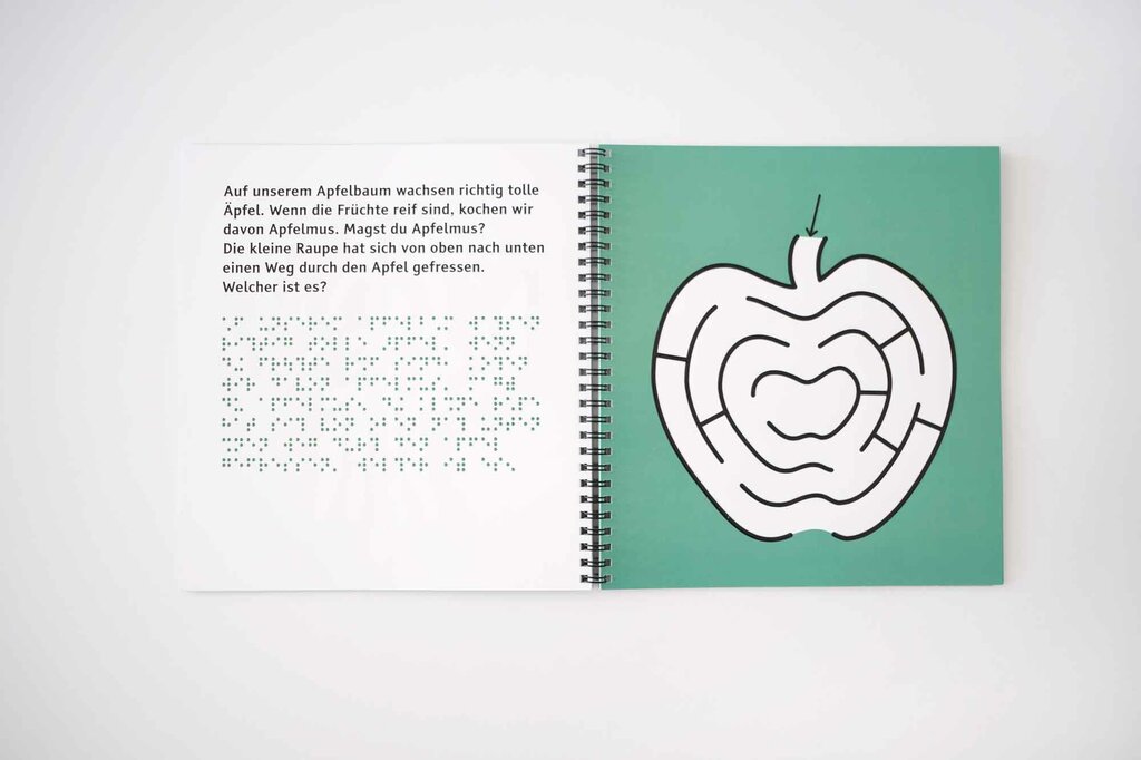 Aufgeschlagenes Ringbuch mit Großdruck, Braille-Lack und Apfel in Lack-Konturen