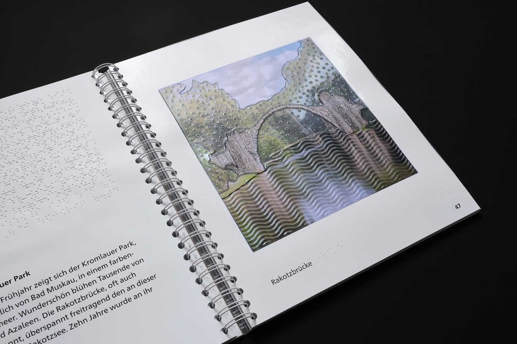 Aufgeklapptes Ringbuch, links Text in Brailleschrift und Großdruck, rechts eine taktile Abbildung der Rakotzbrücke im Kromlauer Park, westlich von Bad Muskau.