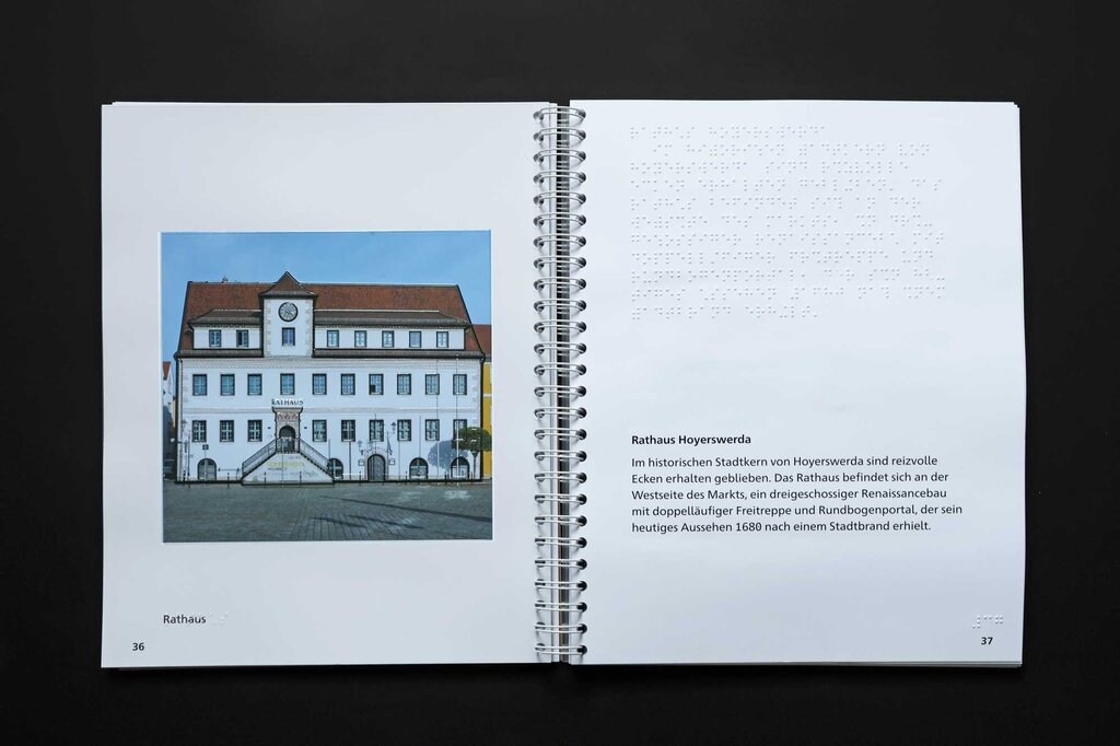 Aufgeklapptes Ringbuch, links eine taktile Abbildung der historischen Fassade des Rathauses in Hoyerswerda, rechts Text in Brailleschrift und Großdruck.