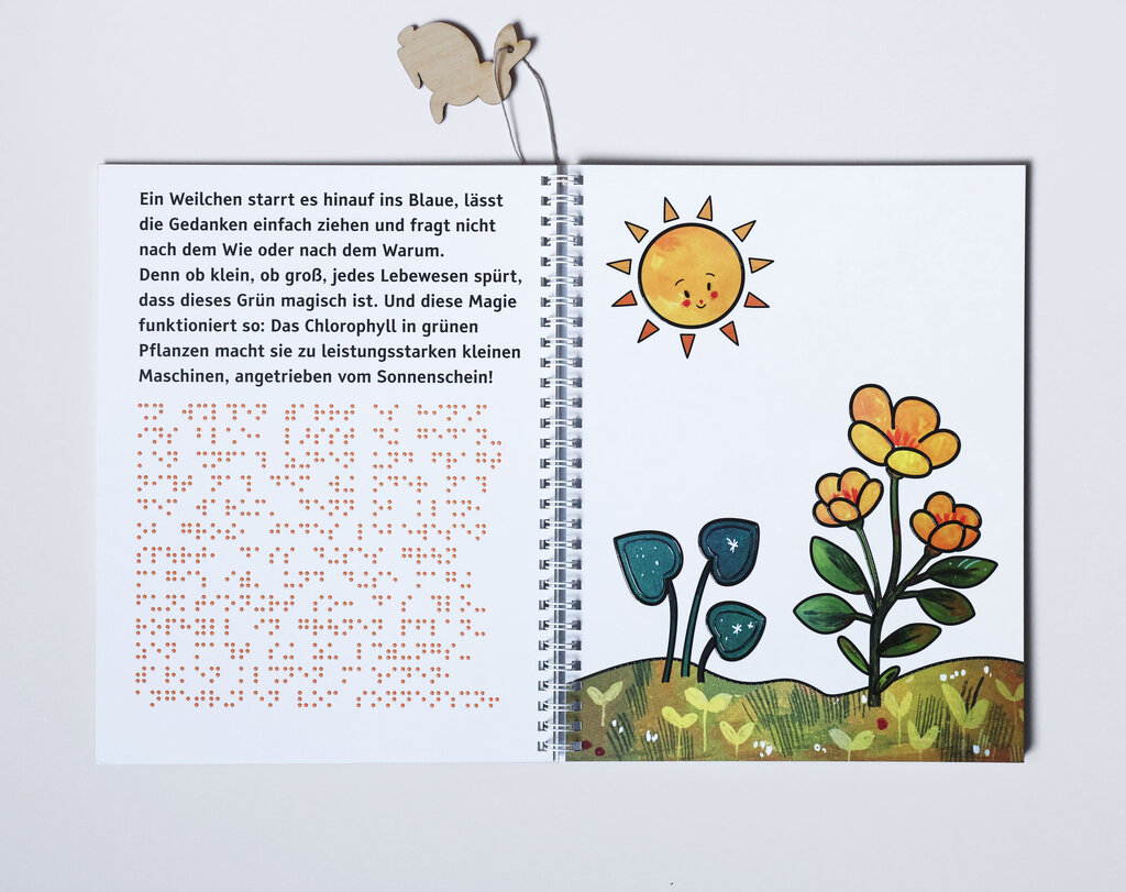 Aufgeklapptes Ringbuch mit Text in Großdruck und Braille-Schrift sowie taktiler Abbildung von zwei verschiedenen Pflanzen unter einer lächelnden Sonne
