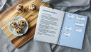 Vergrößerungsansicht: Links Muffins auf einem Holzbrett, darauf liegt ein aufgeschlagenes Ringbuch mit Rezept in Großdruck und Brailleschrift.