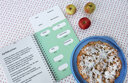 Vergrößerungsansicht: Aufgeschlagenes Backbuch mit Rezept in Großdruck und Brailleschrift, drei Äpfeln und einem runden Kuchen.
