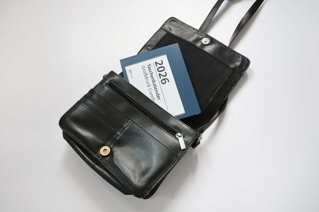 Eine geöffnete schwarze Handtasche mit zur Hälfte eingestecktem  Großdruck-Taschenkalender 2026.