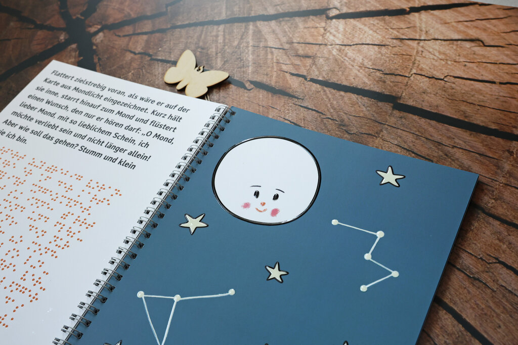 Aufgeschlagenes Ringbuch, links Text in Großdruck und Brailleschrift, rechts eine taktile Abbildung vom Mond, umgeben von Sternen und Sternbildern.