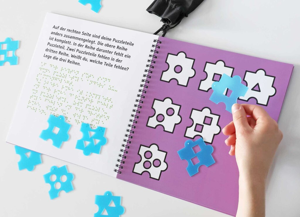 Das Rätselheft »Heute puzzeln wir!« ist aufgeschlagen. Drumherum verteilt liegen Puzzleteile. Eine Hand hält ein Puzzleteil, auf der Suche nach der passenden Form auf der Buchseite.