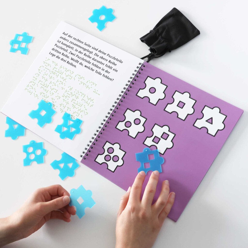 Das Rätselheft ist in Gebrauch zu sehen: Zwei Hände ordnen einzelne Puzzleteile den taktilen Lackkonturen im Rätselheft zu