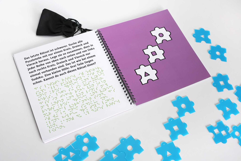 Ein Rätselheft liegt geöffnet auf dem Tisch. Auf der linken Buchseite ist ein Text in Großdruck und Brailleschrift gedruckt, auf der rechten Buchseite befinden sich taktile Lackkonturen in Form von Puzzleteilen. Unter dem Buch liegen verteilt mehrere Puzzleteile.