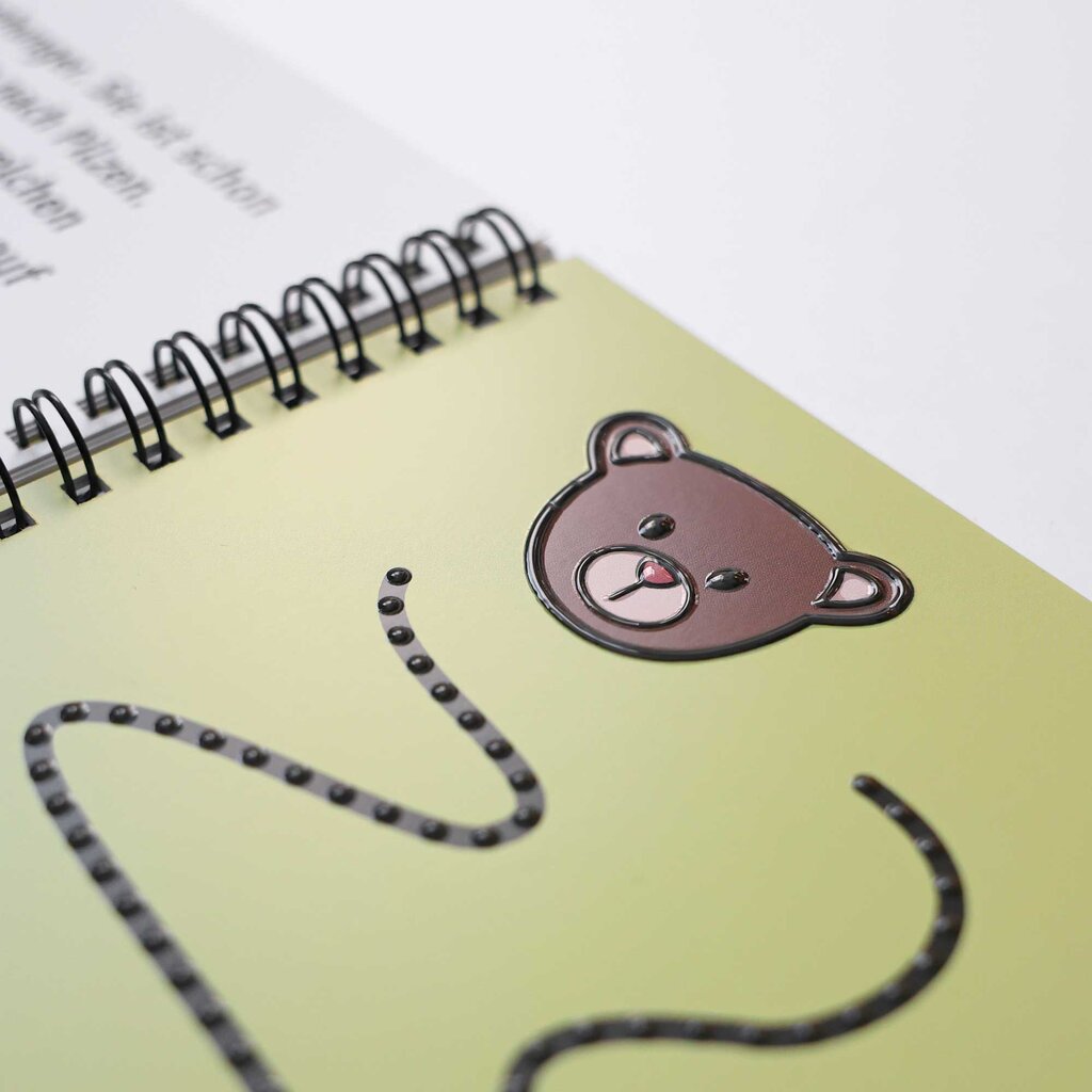 Detailausschnitt einer Seite aus dem Rätselheft »Besuch im Zoo«, abgebildet ist ein taktil gedruckter Bärenkopf, darunter angedeutet eine gepunktete Schlängellinie, ebenfalls tastbar mit Braillelack gedruckt.