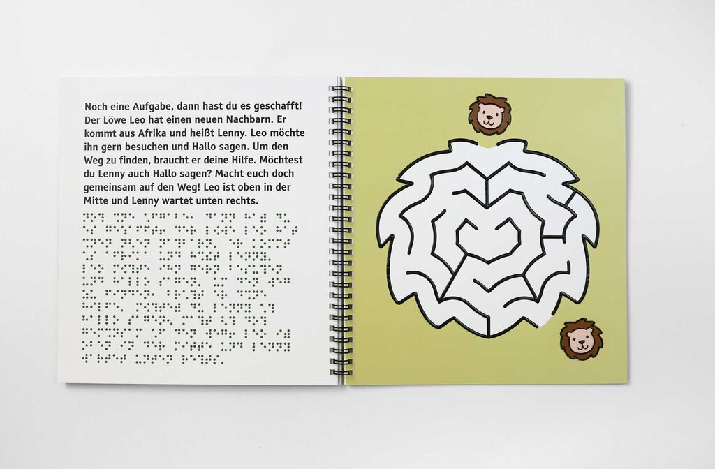 Geöffnetes Rätselheft, links ein Text in Großdruck und Brailleschrift, rechts ein tastbares Labyrinth in Form einer wuscheligen Löwenmähne. Mittig darüber markiert ein Löwenkopf den Eingangspunkt zum Labyrinth. Unten rechts kennzeichnet ein weiterer Löwenkopf den Ausgang des Labyrinths.