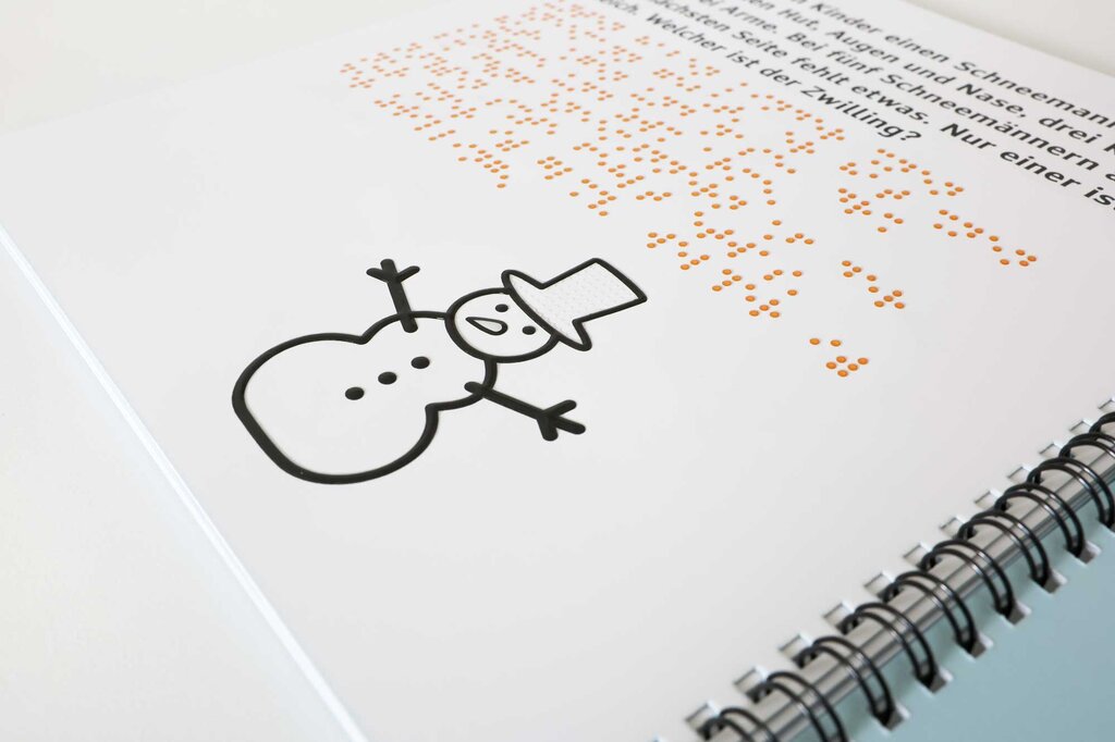 Ein Rätselheft liegt geöffnet auf dem Tisch. Auf der Buchseite ist ein Text in Großdruck und Brailleschrift gedruckt. Unter dem Text befindet sich eine taktile Abbildung von einem Schneemann mit Zylinderhut, kontrastreich gedruckt in fühlbaren Lackkonturen.