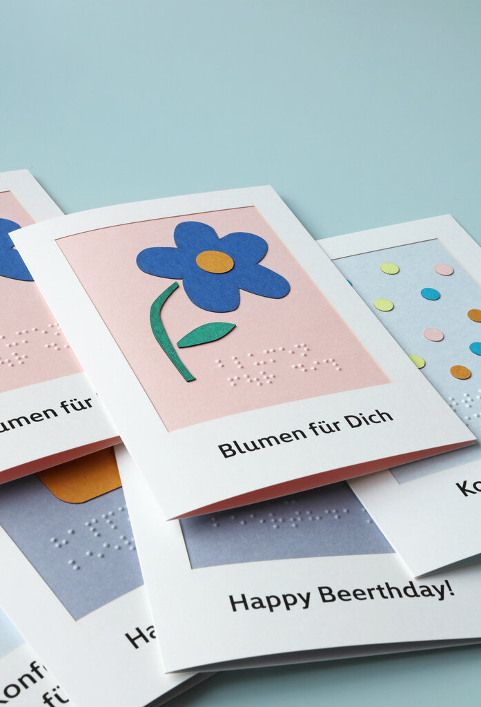 Eine Grußkarte mit tastbarem Blumenmotiv liegt auf einem Stapel Grußkarten.