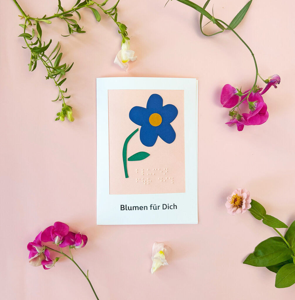 Grußkarte mit tastbarem mit blauer Blüte aus Papier und Text in Brailleschrift und Großdruck auf rosa Hintergrund, umrahmt von verschiedenen Blumen