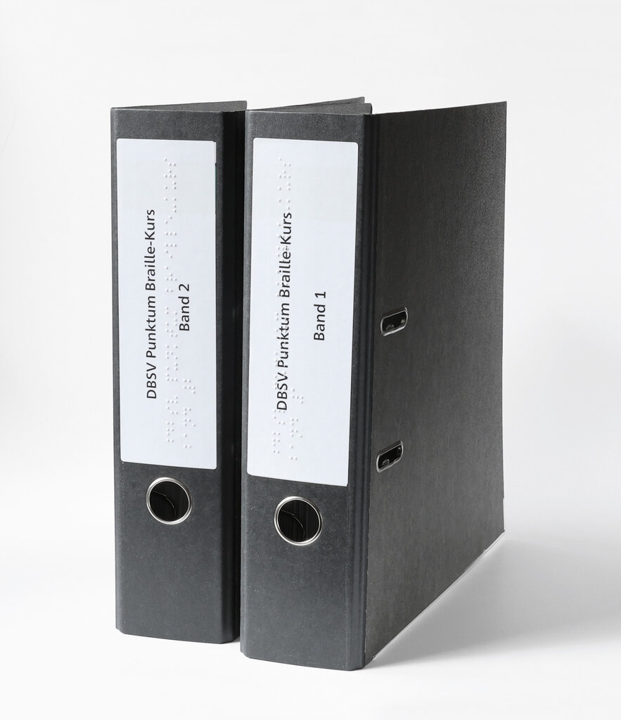 Zwei stehende Ringordner im A4-Format mit Beschriftung in Brailleschrift und Großdruck.