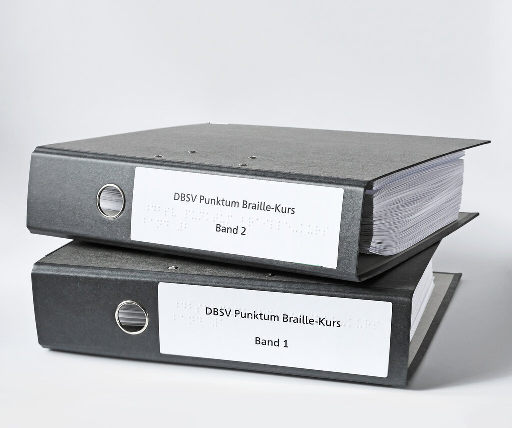 Zwei übereinandergestapelte Ringordner im A4-Format mit Beschriftung in Brailleschrift und Großdruck.