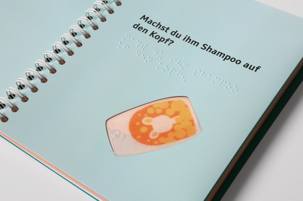 Aufgeschlagene Buchseite, im oberen Bereich ein Text in Großdruck und Brailleschrift, darunter eine tastbare Shampooflasche aus Kunststoff.